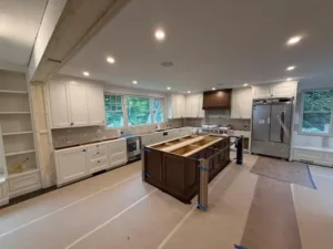 Overview of the entire space showing the custom cabinetry installation in progress, including the island, wood range hood, and a large built-in bookshelf unit.