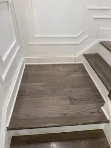 A close-up photograph of the completed staircase landing wood flooring installation. The finished grey-toned oak planks are perfectly fitted into the corner where detailed white wainscoting meets the dark wood stair treads.