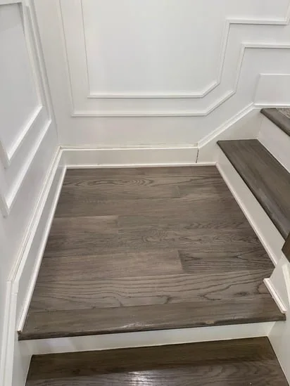 A close-up photograph of the completed staircase landing wood flooring installation. The finished grey-toned oak planks are perfectly fitted into the corner where detailed white wainscoting meets the dark wood stair treads.